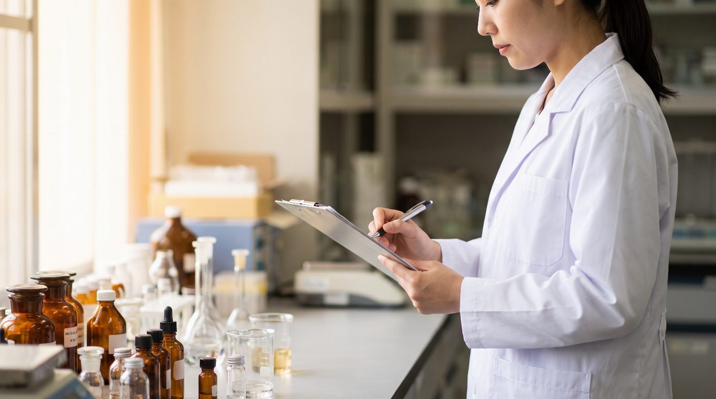Scientist in a lab coat recording notes on a clipboard beside laboratory glassware